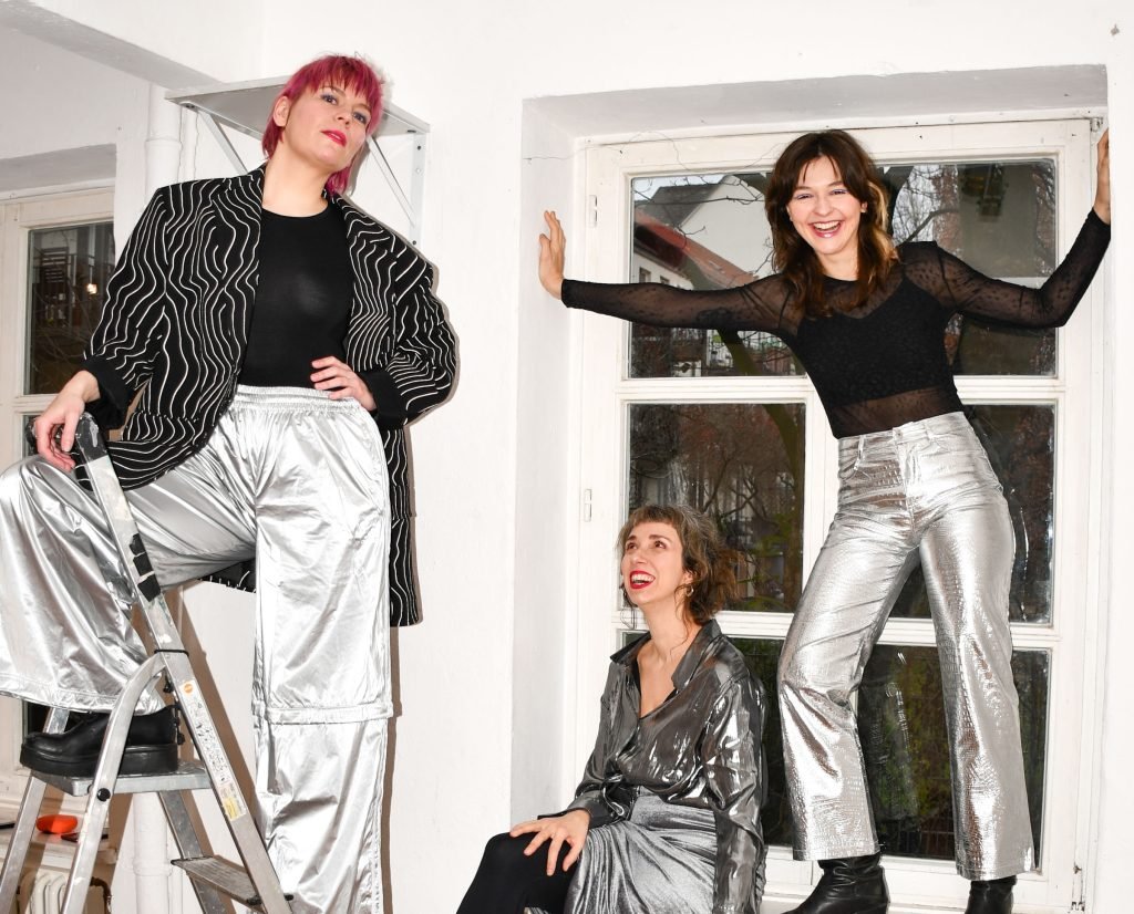 The three members of Bright Future Kollektiv in silver clothes, posing on a ladder, a radiator and a window sill in a white room
