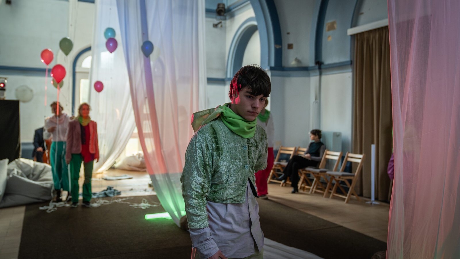 A young performer in a shimmering green jacket and green scarf walks between sheer curtains in the chapel, with balloons, beanbags and seated people behind.
