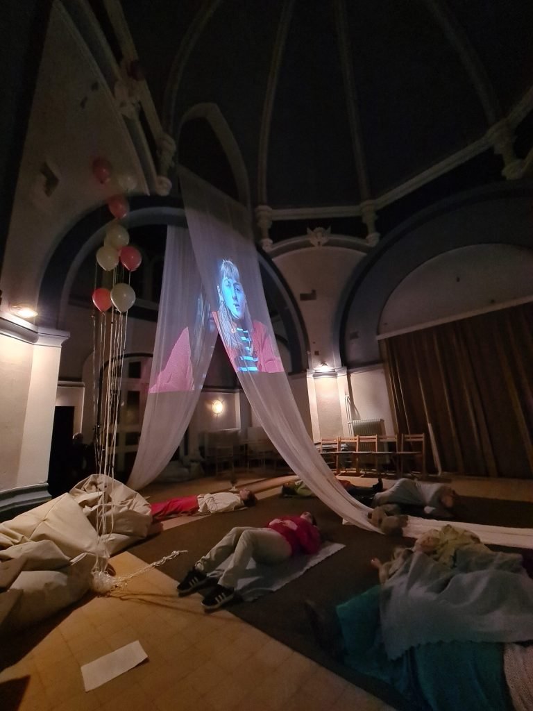 A face is projected onto sheer fabric hanging across the domed chapel, with balloons and stacked chairs around the walls.