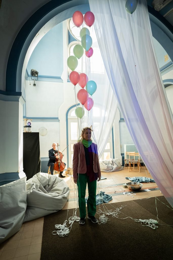 A performer in a pink jacket and green scarf stands under a high arch beside floating balloons and white drapes, while a cellist plays in the background.