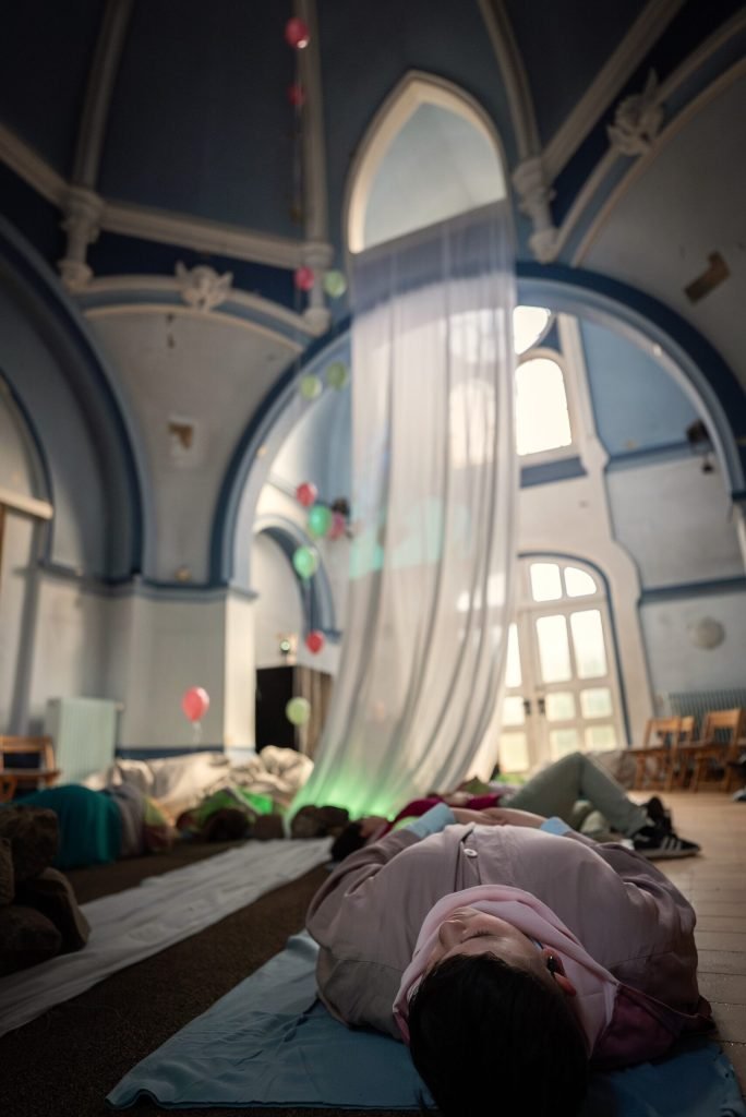 A person lies on a mat in the domed chapel, with long white fabric hanging from the ceiling and balloons around the hall.