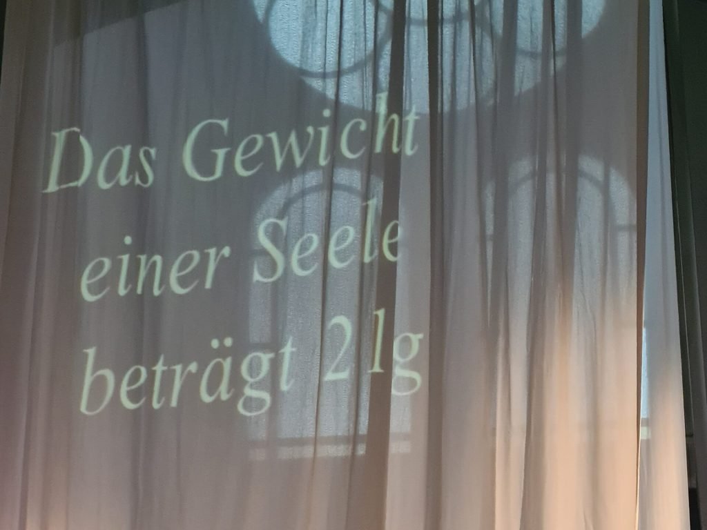 The words "Das Gewicht einer Seele beträgt 21g" (a soul weighs 21 grams) are projected onto a white curtain.