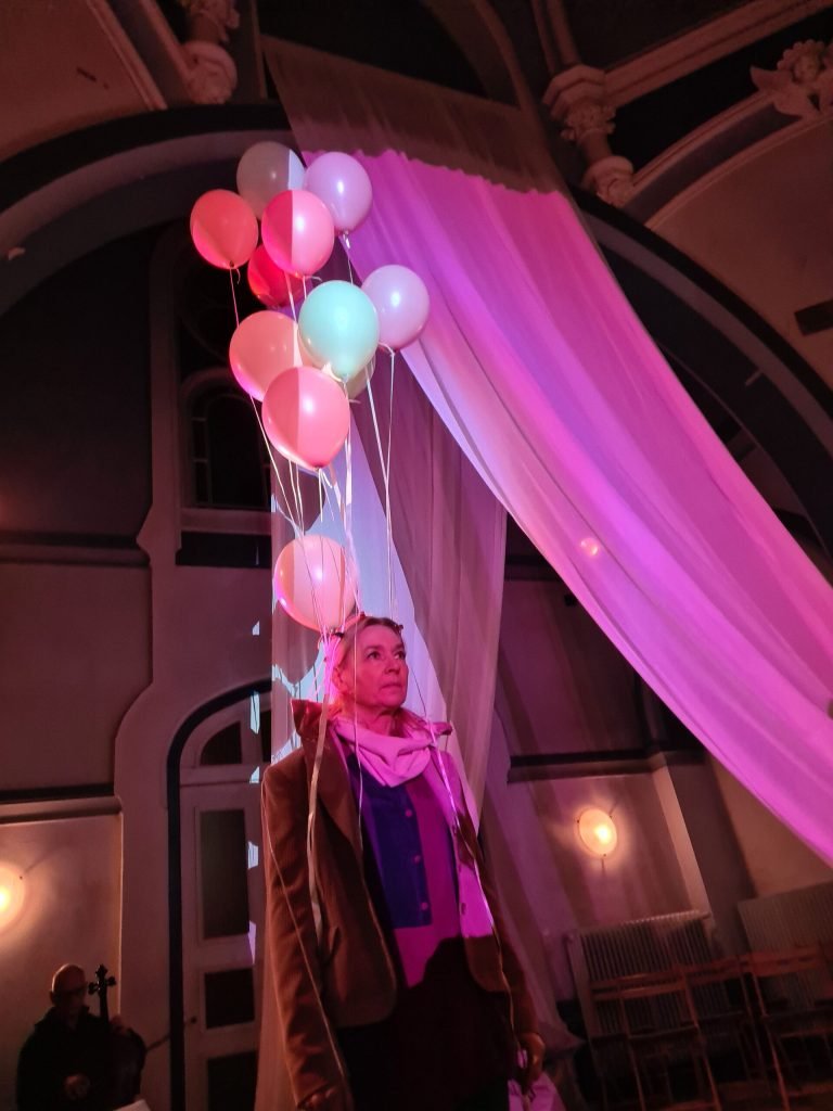 A performer holding a bunch of balloons stands beneath a curtain lit bright pink.