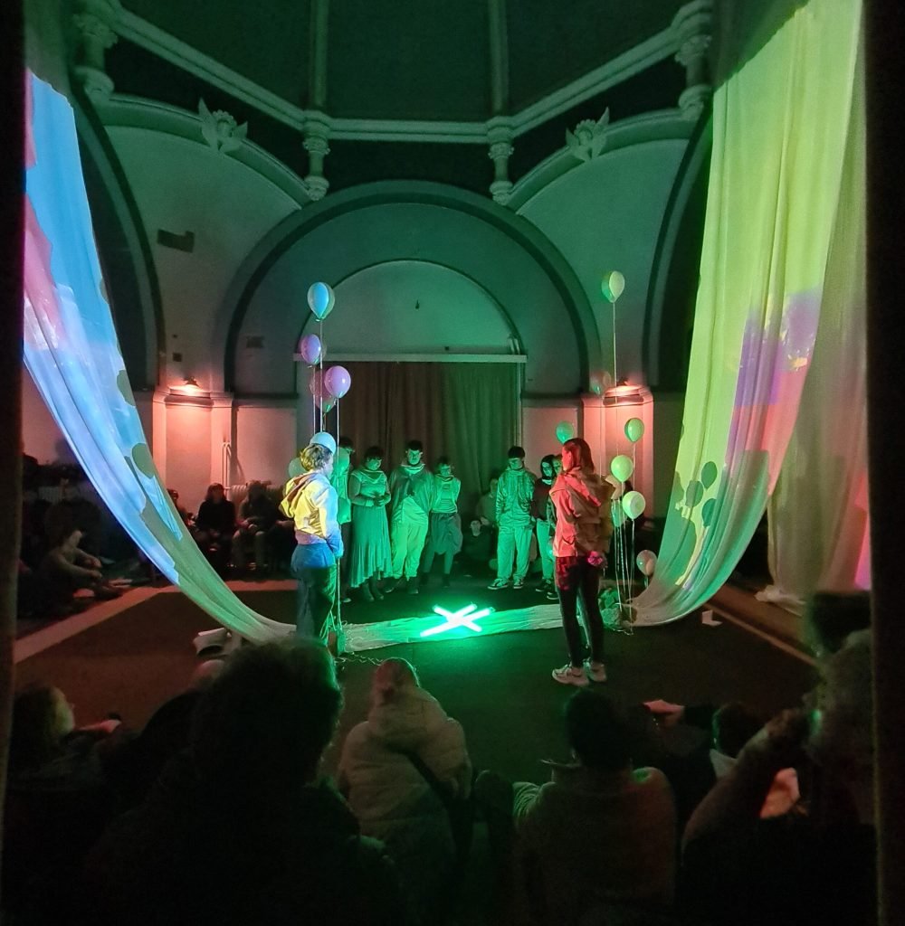 Performers in pale green costumes stand at the far end of the dark chapel, lit green between two glowing curtains, while the audience sits on the floor.