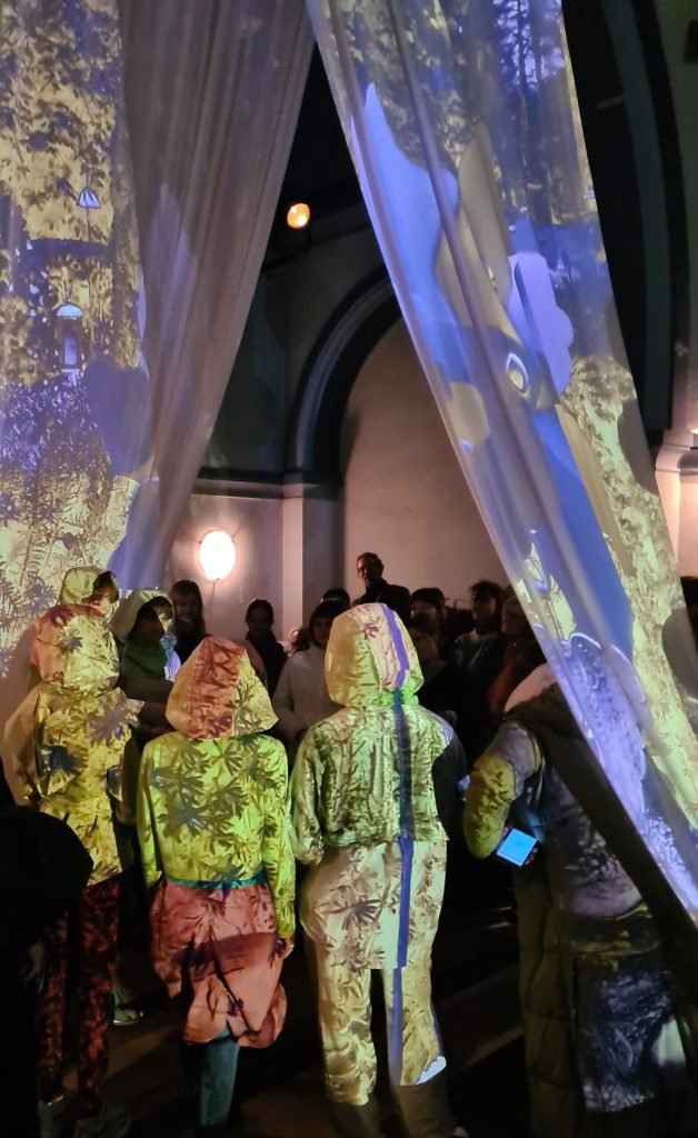 Performers in hooded, patterned costumes stand among the audience under curtains lit with projected leaves.