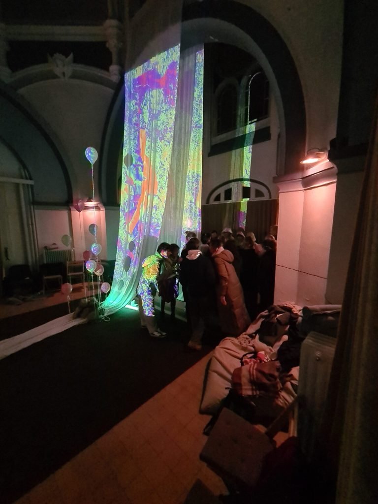 Colourful projections cover a hanging curtain while the audience stands close around it.
